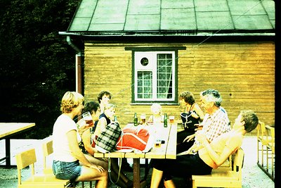 Vintage group dining outdoors at rustic wooden cabin, likely 1970s. Six people seated around a picnic table, enjoying drinks ...