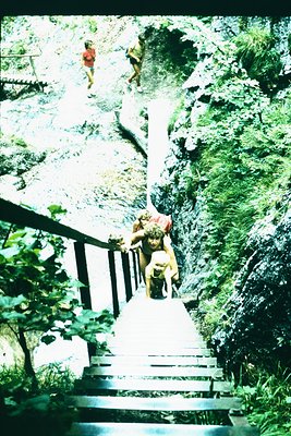 Vintage black-and-white photo of three children descending a concrete staircase beside a cascading waterfall in a lush, fores...