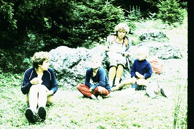 Four individuals in outdoor setting, mid-1970s. Two seated on grass, one kneeling, one standing beside large rock. Casual att...