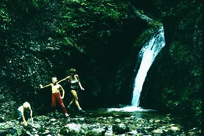 Three individuals explore a lush, rocky waterfall cave, framed by dense greenery. Mid-20th century attire (shorts, tank tops)...