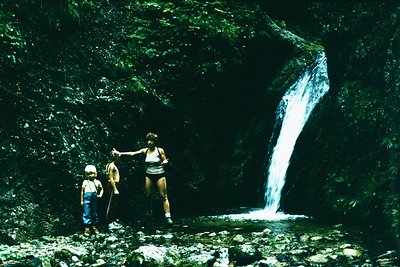 Family poses near a cascading waterfall in a lush, rocky canyon. Mid-20th century attire suggests 1960s–1970s outdoor recreat...