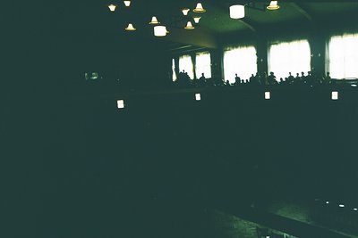 Vintage concert hall interior with tiered seating and suspended industrial-style lamps. Crowd silhouetted against bright stag...