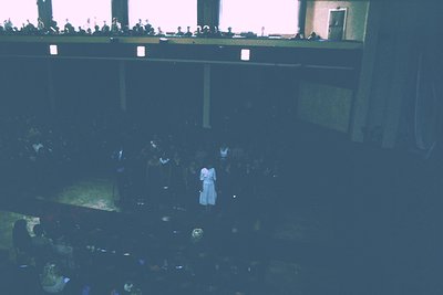 Vintage concert hall interior with stage lighting. Crowd seated in tiered seating, focusing on a performer in a white outfit ...