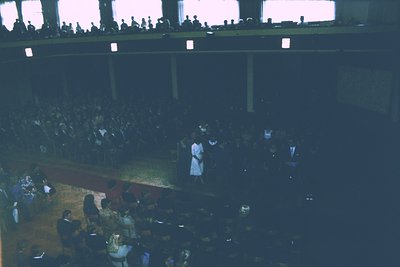 Vintage indoor auditorium shot with tiered seating filled with attendees. Central stage features a podium and floral arrangem...