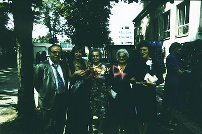 Vintage group portrait in an urban setting, likely 1970s–1980s. Five adults pose outdoors under mature trees, holding flowers...