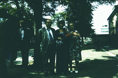 Vintage outdoor portrait featuring three individuals in formal attire: a man in a dark suit with a striped tie, a woman in a ...