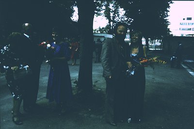 Vintage outdoor gathering with individuals in formal attire, likely a funeral or memorial. A man in a dark suit holds a bouqu...