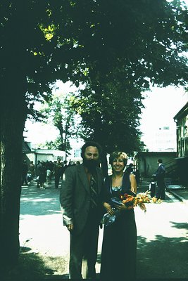Couple in mid-20th century attire (man in suit, woman in floral dress) holding a bouquet of dried flowers, standing under a t...