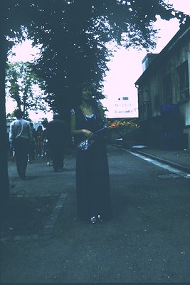 Vintage sepia-toned street scene featuring a woman in a long dress holding a bouquet of flowers, likely from the early-to-mid...