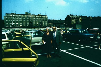 Vintage 1970s urban scene featuring a man and woman in formal attire posing in a parking lot. The woman wears a dark dress wi...