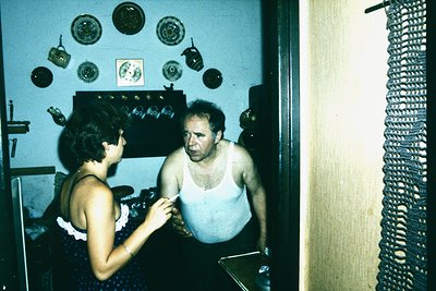 Vintage indoor scene featuring a man and woman in a modestly furnished room, likely mid-20th century. The man, shirtless, sta...