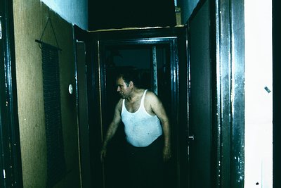 Man in a white tank top exiting dimly lit corridor with peeling paint and industrial lighting fixtures, suggesting institutio...