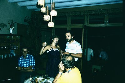 Vintage indoor gathering in a dimly lit, retro-style lounge. Three men and a woman in 1970s fashion—checkered shirt, striped ...