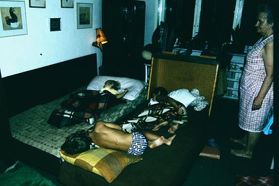 Vintage hotel room interior from the 1960s–70s, featuring two double beds with patterned bedspreads. A woman in a checkered d...