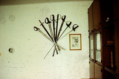 Vintage kitchen utensils arranged in a radial display on a plain wall, including scissors, knives, and a ladle. Framed illust...