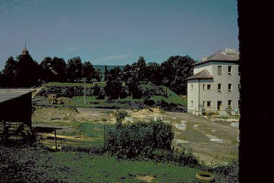 Vintage sepia-toned photo of a rural landscape featuring a two-story brick building with a flat roof and small windows, likel...