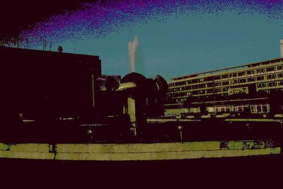 Mid-century Brutalist fountain with geometric concrete base and water jet in foreground. Surrounding buildings feature flat r...