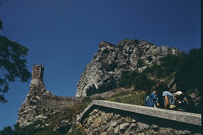 Medieval stone tower perched atop rugged cliffs, connected by a concrete bridge to a rocky hillside. Group of hikers in casua...