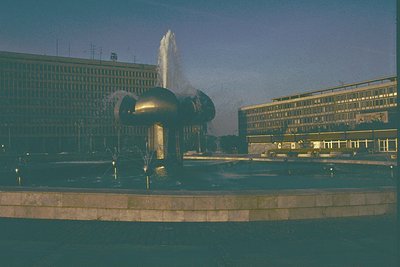 Modernist plaza featuring a striking **1960s-70s Brutalist fountain sculpture** with three curved concrete spouts. Surroundin...