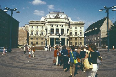 Neoclassical building with grand columns and sculptural details dominates a cobblestone plaza, likely a 1970s European city s...