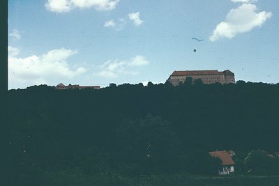 Historic fortress perched atop a forested hill, surrounded by lush greenery. Mid-20th century architectural style with red-ti...