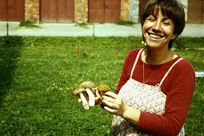 A woman in a red apron and floral blouse holds freshly foraged mushrooms in a lush green garden setting, likely mid-20th cent...