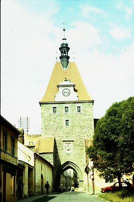 Historic stone gate tower with clock and conical roof, likely a medieval city entrance. Architectural details include arched ...