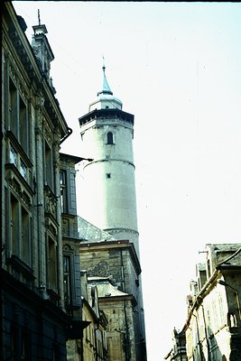 Historic European tower with conical spire flanked by narrow, multi-story buildings. Likely 18th–19th century urban architect...