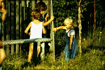 Three children play in a grassy backyard, 1970s. Two girls in overalls and a white sleeveless top lean on a metal fence; a th...