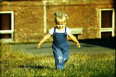 Young girl in 1970s-style overalls runs through grassy courtyard, brick building with small windows in background. Vibrant se...