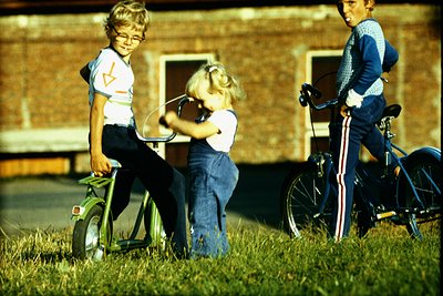 Three children play in a grassy yard, 1970s. Left: boy on a small green tricycle with training wheels, wearing a white T-shir...