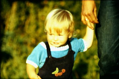 Vintage candid of a young child in overalls and striped shirt, holding an adult’s hand outdoors. Lush greenery and warm light...