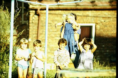 Four children pose under a swing set in a rural backyard, mid-20th century. Stone wall and rustic wooden structure in backgro...