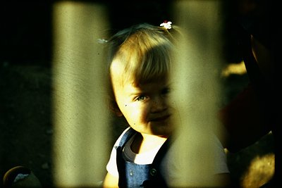 Vintage sepia-toned photo of a young child peering through a window frame, adorned with a floral hair accessory. The child we...
