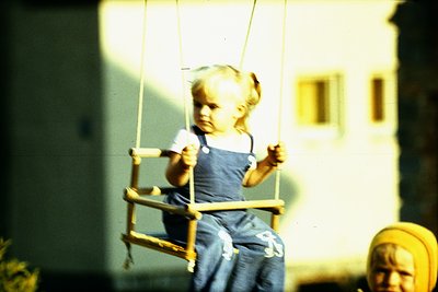 Young child swinging on a wooden swing set in a residential courtyard, mid-20th century. Overalls, pigtails, and vintage sepi...