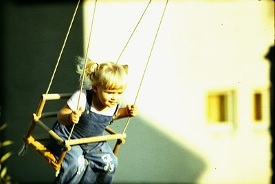 Young child mid-swing on a rope swing in an indoor or semi-covered play area, wearing a white top and denim overalls. Warm li...
