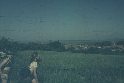 Vintage sepia-toned photo of two women hiking through a lush, grassy field with distant village and church spire. Scratched f...