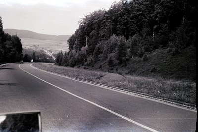 Black-and-white roadside scene featuring a winding asphalt highway flanked by dense greenery and distant rolling hills. Singl...