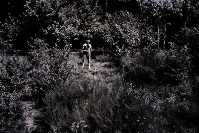 Black-and-white aerial shot of lone figure walking through dense, overgrown forest path. Mid-20th century attire suggests or ...