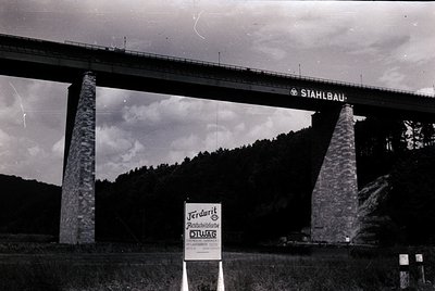 Black-and-white shot of a 1960s German steel bridge ("STAHLBAU") spanning a forested valley. Prominent concrete pillars suppo...