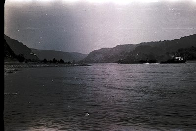 Vintage black-and-white coastal scene featuring calm waters and forested hillsides. Distinctive mist or low clouds obscure di...