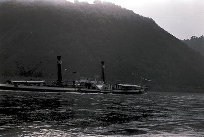 Vintage steamboat navigating mountainous coastline, likely early-to-mid 20th century. Distinctive twin smokestacks and open b...