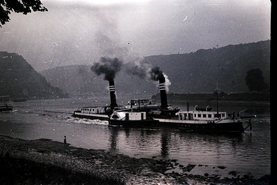 Vintage steamboat navigating a calm river flanked by forested hills, emitting visible smoke from twin smokestacks. Mid-20th c...
