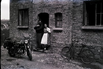 Vintage sepia-toned photo of two individuals posing outside a rustic brick building, likely mid-20th century. The woman wears...