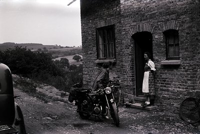 Vintage black-and-white photo of a rural stone house with a woman in a floral dress standing in the doorway, holding a shawl....