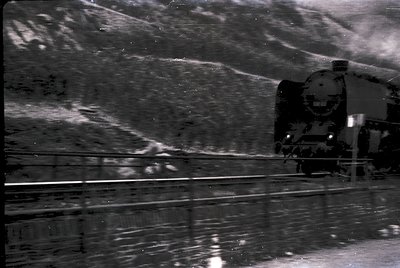 Vintage steam locomotive in motion, captured from inside a moving train car. Reflections on wet tracks suggest recent rain. C...