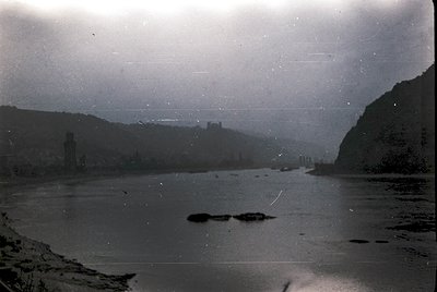 Vintage black-and-white photo of a serene river valley with a ruined castle perched on a hillside. The structure appears medi...