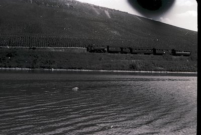 Vintage black-and-white train crossing a low-lying bridge over a calm body of water, with forested hills in the background. M...