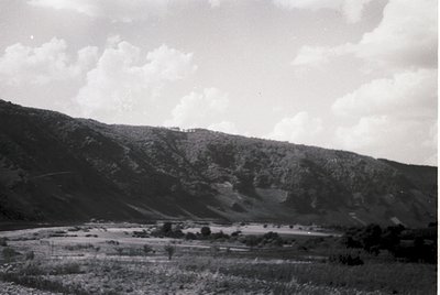 Black-and-white landscape featuring a winding road through a rural valley with rolling hills and sparse vegetation. Mid-20th ...