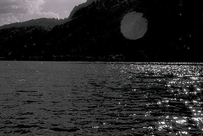 Black-and-white shot of a serene alpine lake reflecting sunlight, framed by forested slopes. The moon’s faint glow contrasts ...